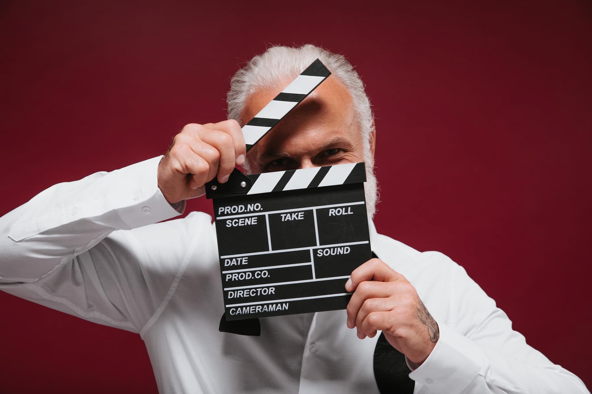 man-in-classic-style-shirt-holds-clapper-white-haired-funny-guy-with-beard-in-light-clothes-posing-on-burgundy-background